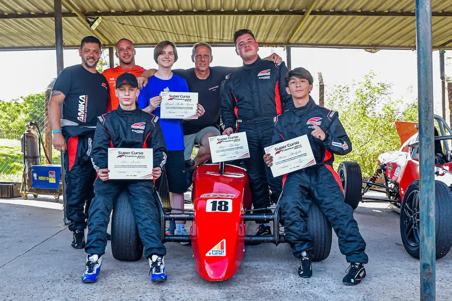 Alunos do Super Curso de Pilotagem da Fórmula Vee Brasil posam com certificados ao lado de carro de corrida monoposto após treinamento em autódromo.