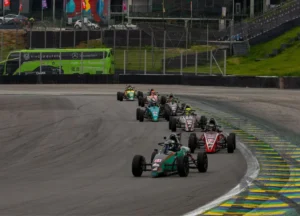 Carros de Fórmula Vee em fila contornando curva de autódromo durante corrida, com pilotos disputando posição em alta velocidade