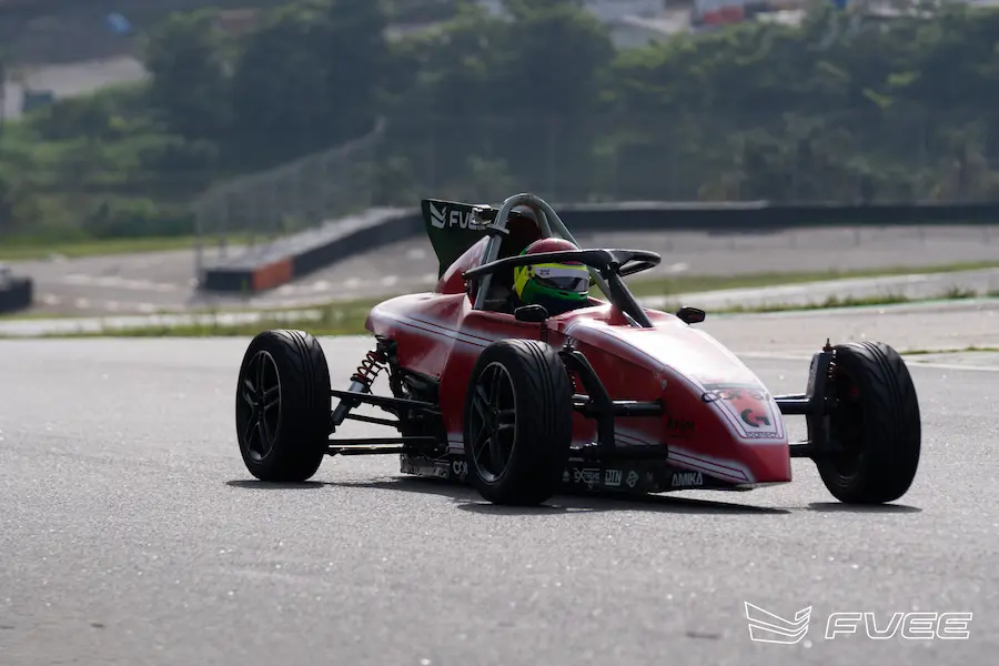 Carro de Fórmula Vee vermelho em ação durante treino em autódromo, com piloto equipado acelerando em pista profissional. A imagem destaca a dinâmica do carro monoposto em curva, transmitindo velocidade, controle e experiência real no automobilismo.