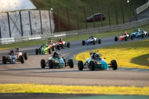 Grid de carros da Fórmula Vee percorrendo uma curva de autódromo durante uma corrida, com vários monopostos disputando posições em pista asfaltada, cercada por áreas gramadas e estruturas de proteção. A imagem destaca a competitividade e a dinâmica dos campeonatos de automobilismo.