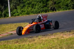 Carro da Fórmula Vee Brasil em ação durante treino em autódromo, contornando uma curva de alta velocidade. O monoposto laranja e preto percorre o traçado asfaltado cercado por área gramada e vegetação ao fundo, destacando a experiência de pilotagem em ambiente profissional de automobilismo.