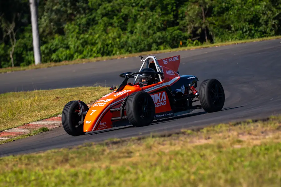 Carro da Fórmula Vee Brasil em ação durante treino em autódromo, contornando uma curva de alta velocidade. O monoposto laranja e preto percorre o traçado asfaltado cercado por área gramada e vegetação ao fundo, destacando a experiência de pilotagem em ambiente profissional de automobilismo.