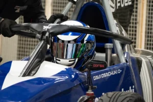 Piloto de Fórmula Vee posicionado no cockpit do carro de corrida durante preparação para entrada na pista, utilizando capacete completo e equipamentos de segurança em ambiente de autódromo.