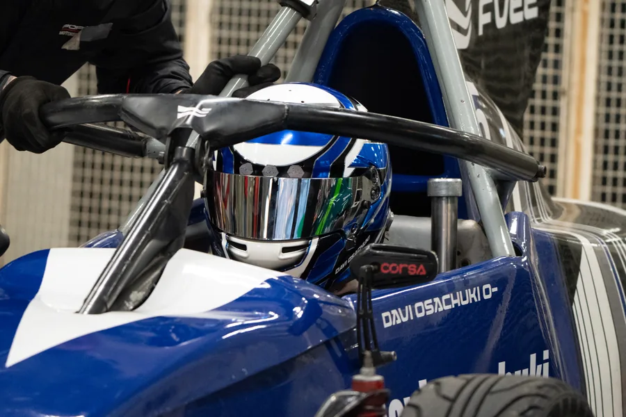 Piloto de Fórmula Vee posicionado no cockpit do carro de corrida durante preparação para entrada na pista, utilizando capacete completo e equipamentos de segurança em ambiente de autódromo.