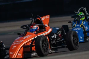 Dois carros de Fórmula Vee em uma pista de corrida, com pilotos equipados e o carro laranja em destaque no primeiro plano.