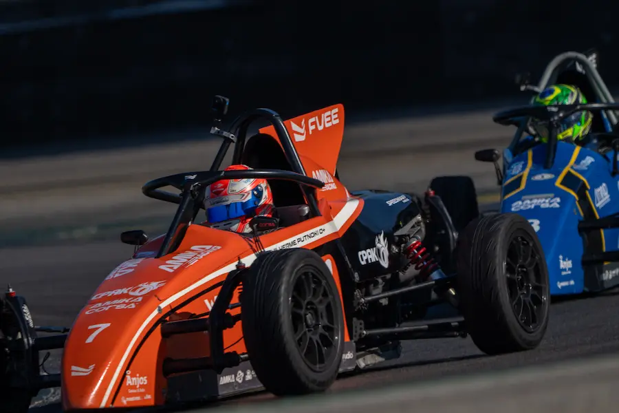 Dois carros de Fórmula Vee em uma pista de corrida, com pilotos equipados e o carro laranja em destaque no primeiro plano.