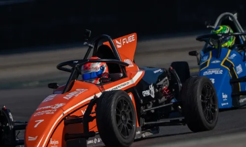 Dois carros de Fórmula Vee em uma pista de corrida, com pilotos equipados e o carro laranja em destaque no primeiro plano.