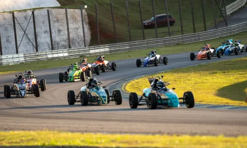 Grid de carros da Fórmula Vee percorrendo uma curva de autódromo durante uma corrida, com vários monopostos disputando posições em pista asfaltada, cercada por áreas gramadas e estruturas de proteção. A imagem destaca a competitividade e a dinâmica dos campeonatos de automobilismo.