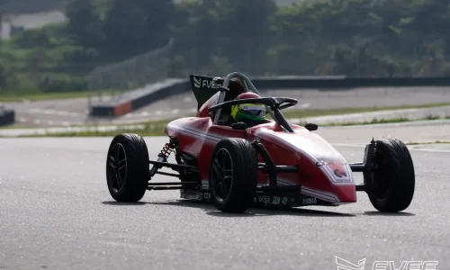 Carro de Fórmula Vee vermelho em ação durante treino em autódromo, com piloto equipado acelerando em pista profissional. A imagem destaca a dinâmica do carro monoposto em curva, transmitindo velocidade, controle e experiência real no automobilismo.