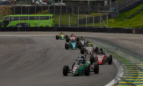 Carros de Fórmula Vee em fila contornando curva de autódromo durante corrida, com pilotos disputando posição em alta velocidade