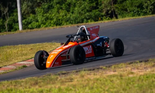 Carro da Fórmula Vee Brasil em ação durante treino em autódromo, contornando uma curva de alta velocidade. O monoposto laranja e preto percorre o traçado asfaltado cercado por área gramada e vegetação ao fundo, destacando a experiência de pilotagem em ambiente profissional de automobilismo.