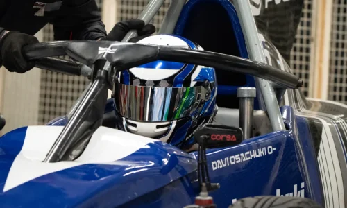 Piloto de Fórmula Vee posicionado no cockpit do carro de corrida durante preparação para entrada na pista, utilizando capacete completo e equipamentos de segurança em ambiente de autódromo.